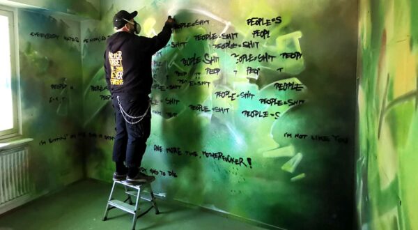 Person writing texts on a greenish wall with black spraypaint.